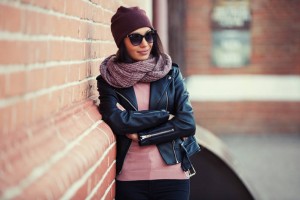 Young fashion woman in sunglasses leaning on brick wall Stylish female model wearing black leather jacket and knitted scarf